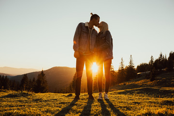 Happy couple hugging and kissing at sunset with amazing mountain view. Warm evening, sun light, couple hug together and close to a pine forest. 
Sun between the legs of a couple. Copy space.