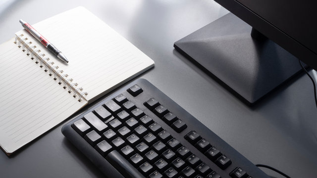 Selective focus at black keyboard with part of computer monitor, ballpoint pen and spiral note pad on gray tabletop with sunlight and shadow on surface