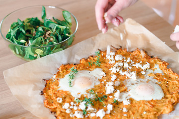 Hand dropping sheep milk cheese on potato pancake with egg and green salad on wooden plate