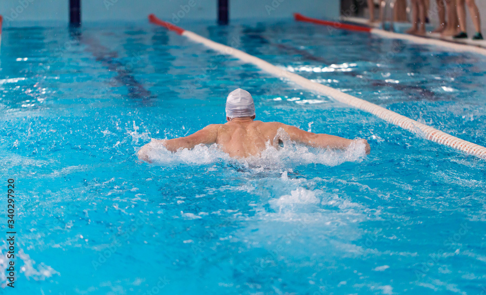 Wall mural Swimming pool athlete training indoors for professional competition. Teenager swimmer wearing white swimming cap performing the butterfly stroke