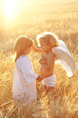 A baby with wings touches a second by the hair in a field at sunset