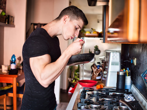 Young Male Bodybuilder Cooking In Kitchen At Home, Eating From Pan While Preparing Food, Looking Down At It