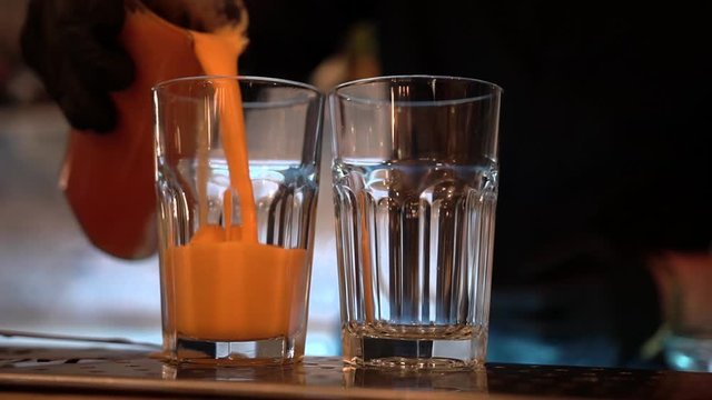 A bartender silhouette fills two transparent glasses with orange juice smoothie on a bar background, close-up.