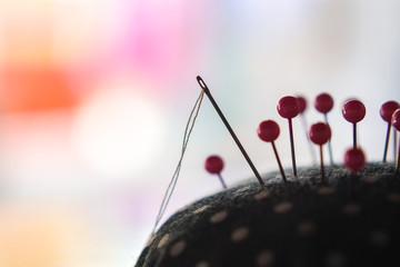 Needle with thread and pink pins on pincushion for handmade sewing cloth with bokeh and blurred background close-up.