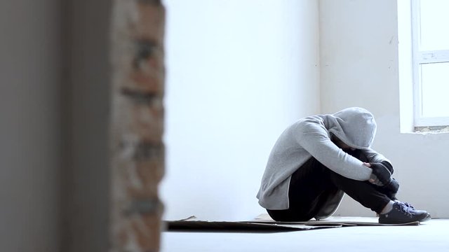 Nervous guy in hood is sitting on cardboard on floor and suffering in abandoned building. Homeless sad man lost everything because of addiction. Social problems in society. Loneliness and depression.