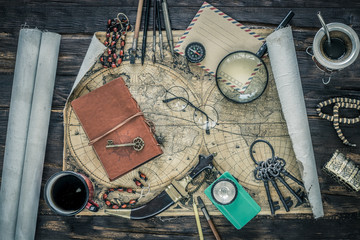 Preparing for travel.  Great adventure planning concept.  Retro style.  Old maps, expedition accessories.  Notebook, magnifier, flashlight, compass, tea cup, treasure chest and key on a wooden bench