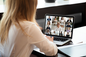 View over businesswoman shoulder sit at workplace desk looks at pc screen during group videocall with different age and ethnicity colleagues using application and webcam, distant communication concept - obrazy, fototapety, plakaty