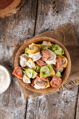 Top view on raw semi-finished multi-colored dumplings in a bowl on the wooden table with sour cream, vertical