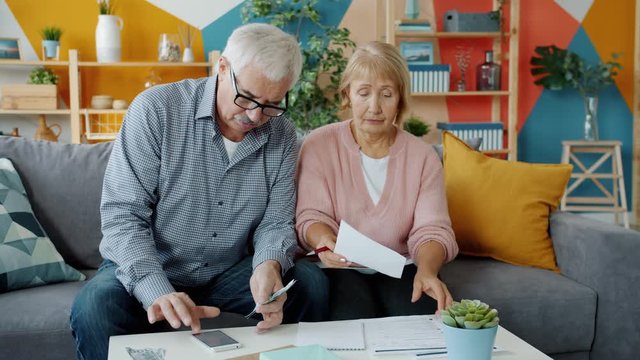 Serious senior couple is dealing with bills reading financial papers and counting money in apartment. Household, family and modern lifestyle concept.