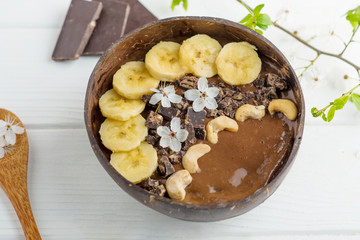 chocolate smoothie, banana with cashew in a coconut bowl, bamboo spoon on a white table with flowers. vegan breakfast and healthy food