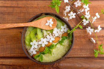 kiwi smoothie, spinach, banana in a coconut bowl, bamboo spoon on a wooden table with flowers. vegan breakfast and healthy food