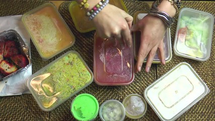 Closeup over head POV shot of a woman’s hands removing lids from plastic containers from an Indian takeout / takeaway meal, on wicker table mats.