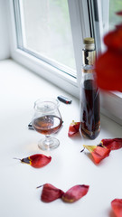 red wine and rose petals. cognac in a bottle and in a glass, red flower petals on a white window. loneliness and broken heart concept. living grief. celebrating the end of a hard day