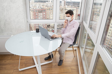 A guy in a protective face mask is talking on the phone and working at a computer from home during a virus epidemic. Quarantine during viral infection