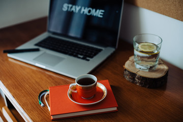 Cozy workplace of a freelancer: opened laptop with words in English "Stay home", red notebook, glass of water with lemon and red cup of espresso. Remote work or work from home during Covid-19 pandemic
