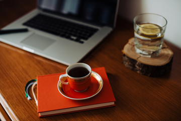 Morning of a freelancer, cup of coffee, glass of water with lemon, opened laptop, notebook on the desk. Work from home concept