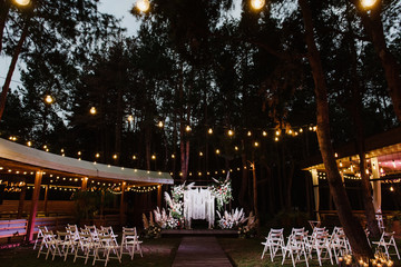The boho-style wedding arch is decorated with flowers and light bulbs. Evening wedding ceremony.