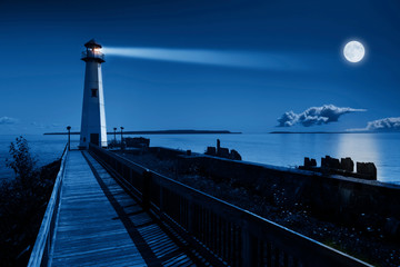 This is a beautiful photo illustration of a dramatic night time scene of a blue moonrise in a clear sky on a ocean pier with a brightly lit lighthouse beacon and calm ocean waters.