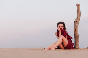Fototapeta na wymiar Girl sitting in the desert in the wind in a red dress near an old tree