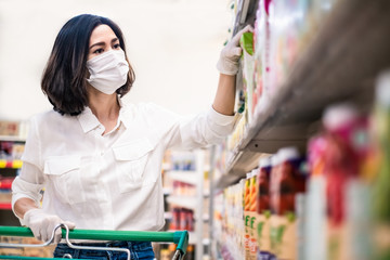 Fototapeta na wymiar Asian woman wearing face mask and rubber glove push shopping cart in suppermarket departmentstore. Girl choosing, looking grocery things to buy at shelf during coronavirus crisis or covid19 outbreak.