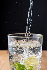 Top view of glass of water with falling water, mint, lemon and bubbles, vertically, on wooden table and black background, vertically, with copy space