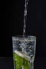 Close-up of water falling into glass cup with lemon and mint, on black background in vertical, with copy space