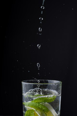 Fototapeta na wymiar Close-up of water drops falling into glass cup with lemon and mint, on black background in vertical, with copy space