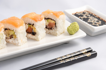 Sushi roll with pink salmon wrap, grilled salmon, Philadelphia cheese and avocado. White container and black chopsticks.