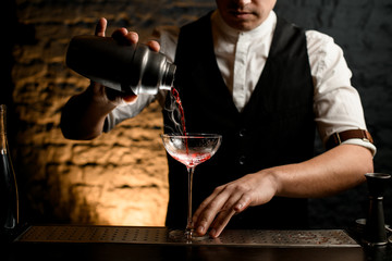 Close-up professional bartender pouring cocktail from stainless shaker into glass