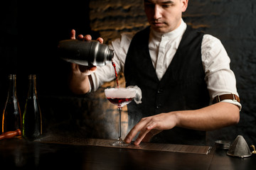 professional man bartender carefully pouring cocktail from stainless shaker into smoky glass
