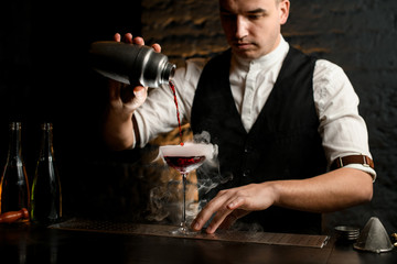 Man at bar carefully pouring cocktail from stainless shaker into smoky glass