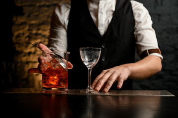 Close-up barman holds glassy mixing cup with strainer full of cold alcoholic drink with pieces of ice.