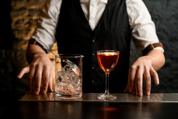 Close-up. Clear glassy mixing cup with strainer and glass full of cold alcoholic drink stand on bar