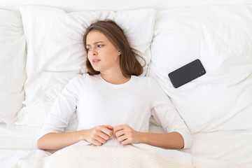 Young female put away her smartphone after breakup with lover, lying in bed in morning, looking helpless, stressed and lonely
