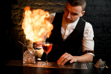 Close-up. Barman actively sprinkles on glass with cocktail and make fire.