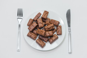 Chocolate bars on white plate with fork and knife on white background. Unhealthy high-calorie diet