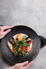 Salad with arugula and seafood on a plate with hands. Salmon, mussels, shrimp, prawn, white fish and grapefruit on a light grey background. Top view, flatlay. Background image, copy space