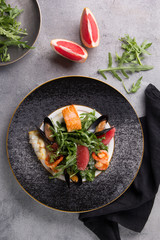 Salad with arugula and seafood on a plate with black cloth. Salmon, mussels, shrimp, prawn, white fish and grapefruit on a light grey background. Top view, flatlay. Background image, copy space