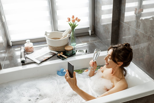 Young woman taking a bath, drinking sparkling wine, talking on phone, lying in bathtub with foam near the window at home