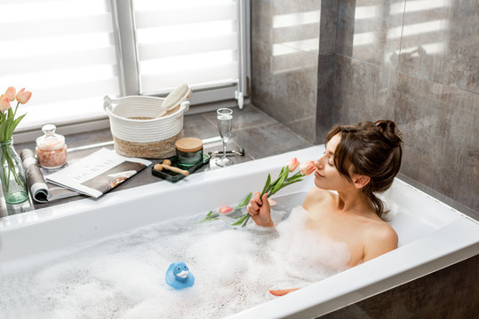 Young woman taking a bath, lying in the bathtub with flowers and rubber duck, relaxing in the bathroom at home