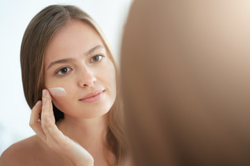 Headshot of young lady standing close to bathroom mirror, touching her skin, applying moisturizing cream, smiling peacefully, enjoying beauty routine
