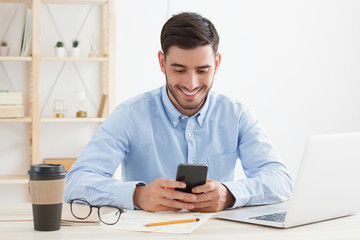 Male office worker dressed in blue shirt enjoying break from work, scrolling news feed on phone, having fun at content, smiling happily