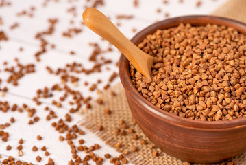 Close-up of buckwheat with a spoon in a ceramic dish on a napkin and scattered on a white wooden table.