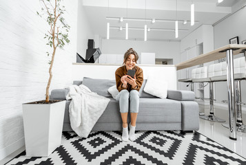 Young relaxed woman sitting with mobile phone on the couch at the living room at home, wide view on...