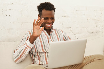 Happy handsome young African male sitting on couch with laptop staying connected with family via...