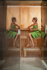 Young and relaxed woman covered with green towel sitting in a small Finnish sauna