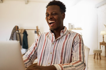 Happy cheerful young African male relaxing at home surfing internet on laptop, smiling broadly,...