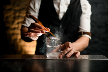 close-up hand of bartender professionally holds tweezers with ice and puts it in steaming glass