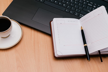 Notebook with a pen,laptop,white cup of coffee and on the wooden table.Concept work from home.Remote work due to the coronavirus pandemic.Top view,copy space for text