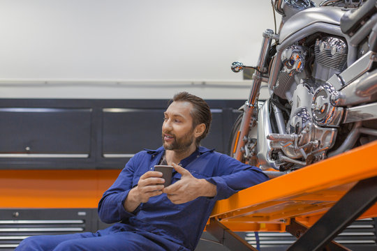 A smiling man in the workshop sits near a motorcycle and drinks coffee.